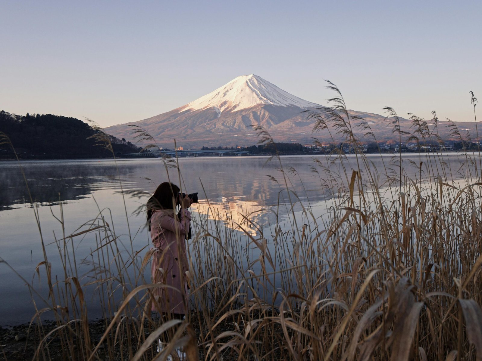 A woman captures the scenic beauty of Mount Fuji reflecting on a lake at sunset.