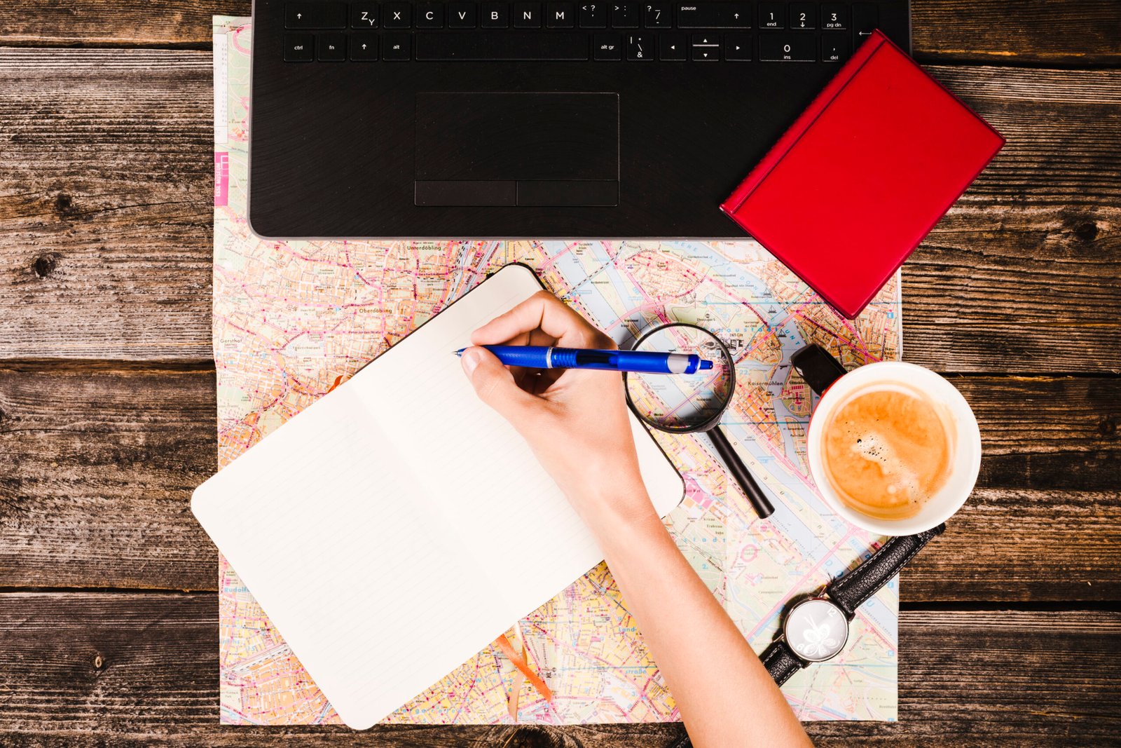 A person organizing a travel budget with a laptop, notebook, and coffee cup on a wooden table.
