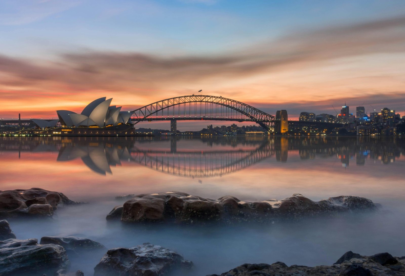 Stunning sunset view of Sydney Opera House and Harbor Bridge reflecting over calm waters.