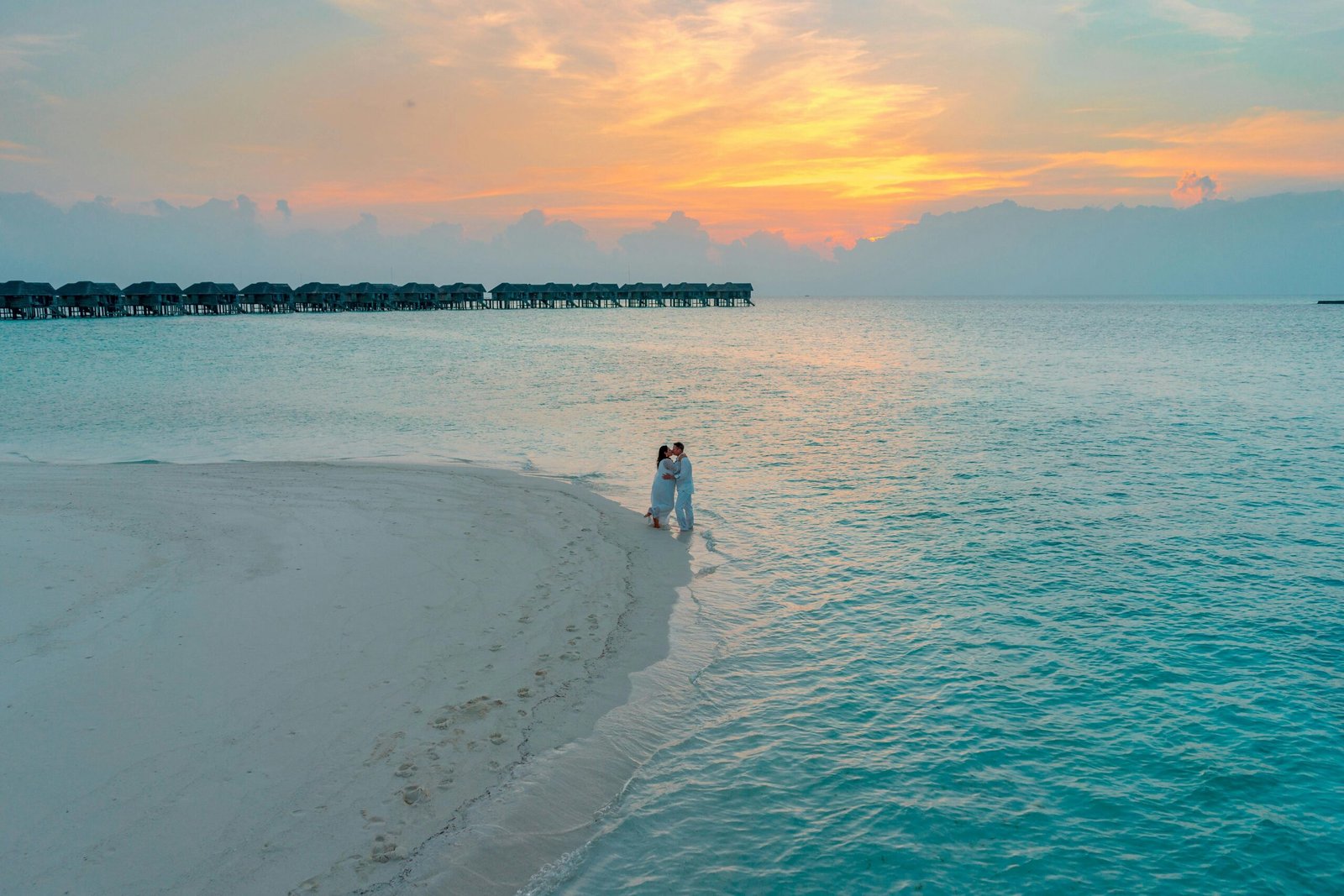 Aerial view of a couple embracing on a sandy beach at sunset with overwater bungalows in the background.