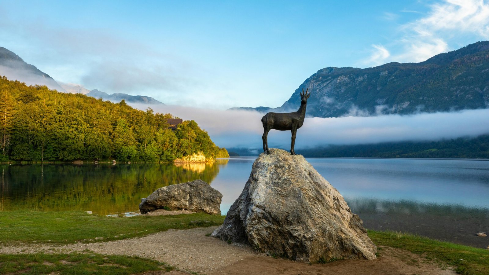Bronze animal sculpture on rock by Lake Bohinj with misty mountain backdrop at sunrise.