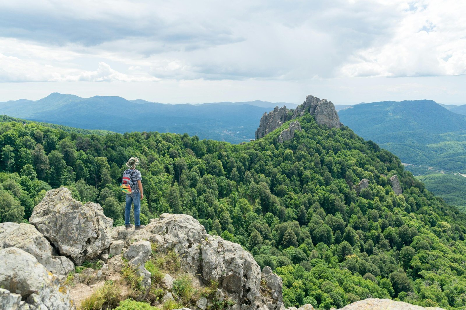Solo hiker stands on rocky mountain peak overlooking lush green valley and rugged hills.