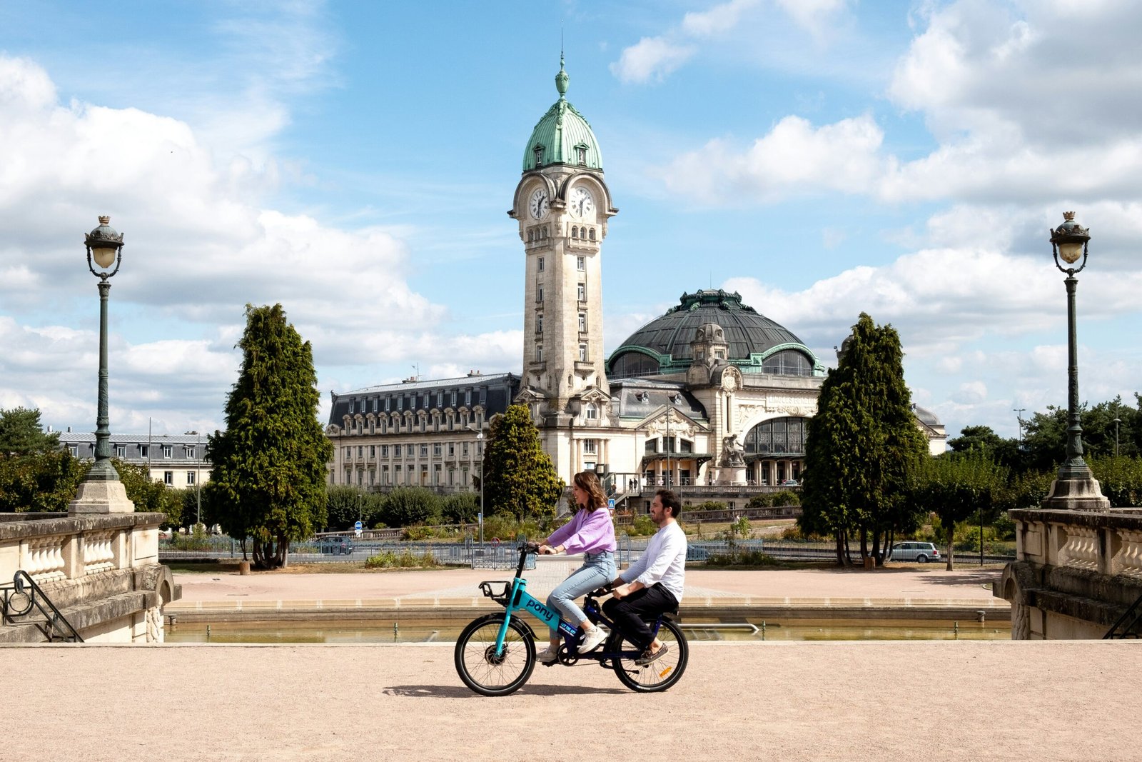 A traveler using a bike-sharing service in a European city, with green spaces and a clear summer sky.
