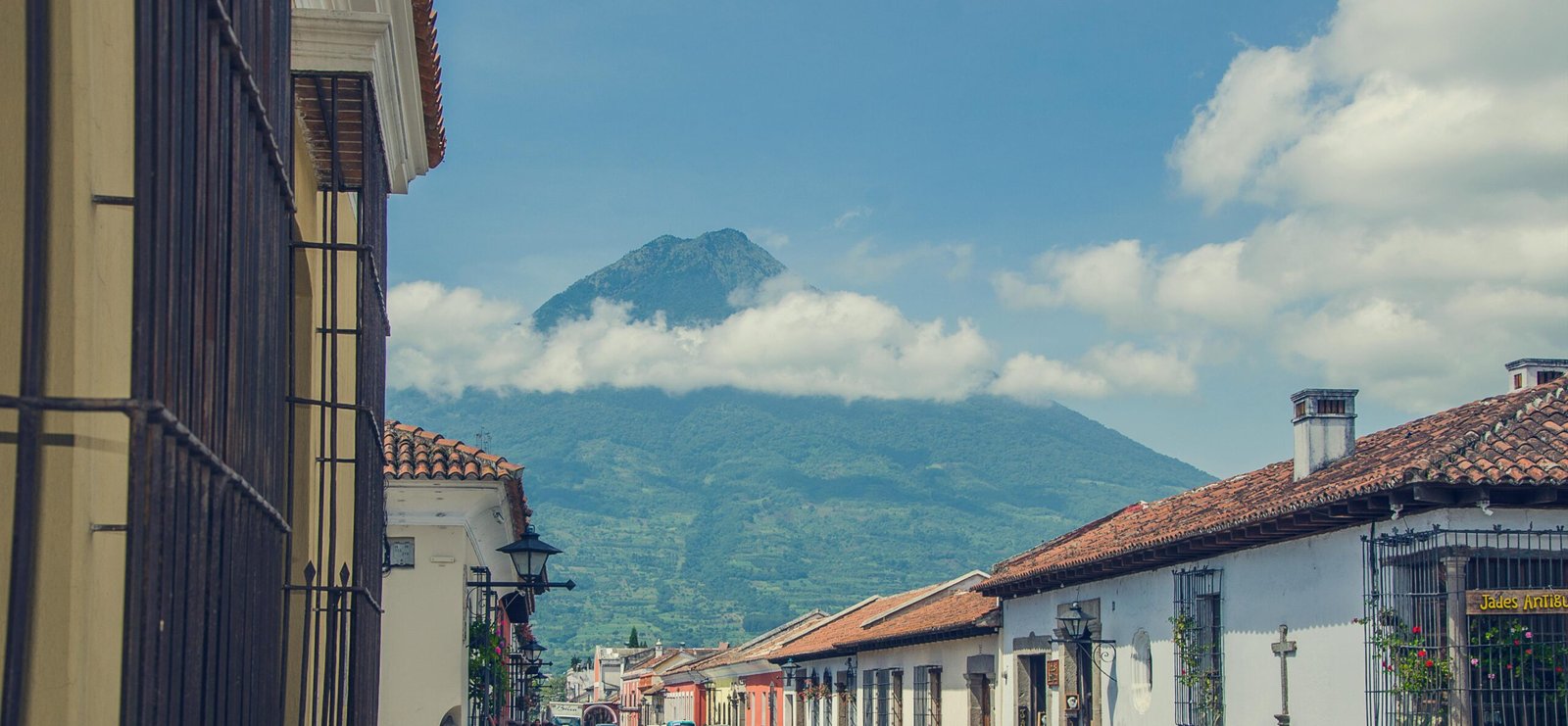 A colorful, colonial street in Antigua, Guatemala, with cobblestones and the majestic Volcán de Agua visible in the background.