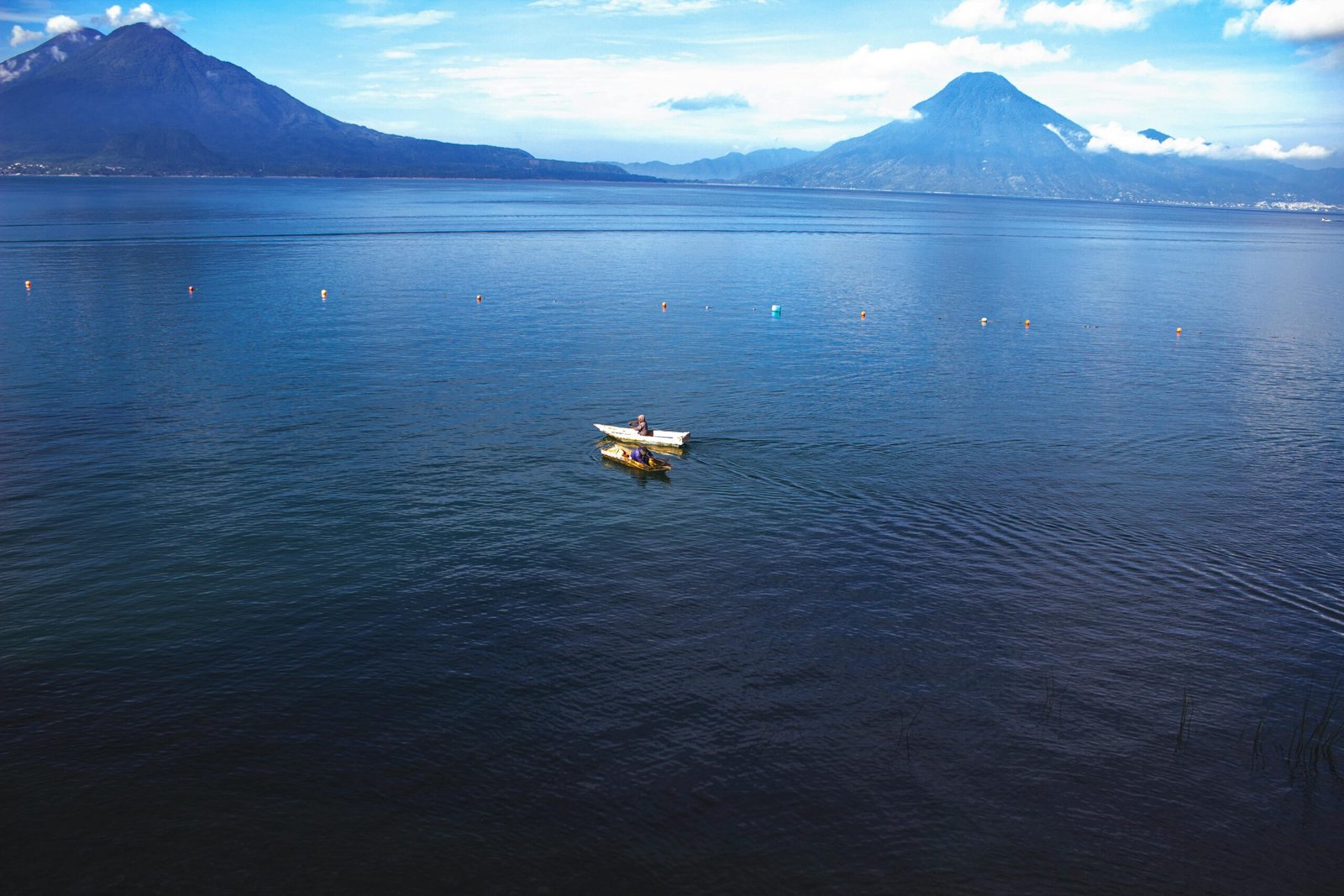 A classic, breathtaking shot of a traditional wooden boat (lancha) on the deep blue waters of Lake Atitlán, with the three majestic volcanoes rising dramatically in the background.